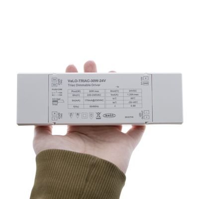 Henkilö, jolla on "VaLO-TRIAC-30W-24V" LED-ohjain, jonka tekniset tiedot ovat valkoisella pinnalla, ihanteellinen LED-valaistusjärjestelmiin.
